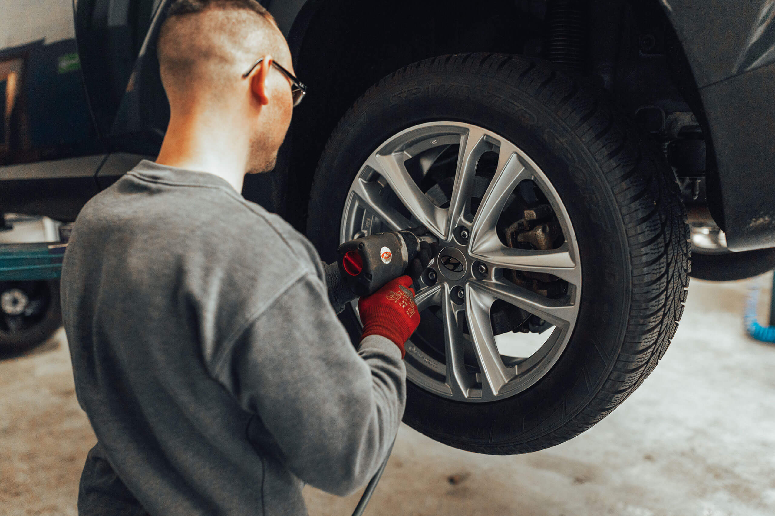 Ein Mann in grauem Pullover und roten Handschuhen befestigt mit einem Schraubwerkzeug ein Rad an einem aufgebockten Auto in einer Werkstatt. Das Auto ist teilweise sichtbar und der Boden ist betoniert.