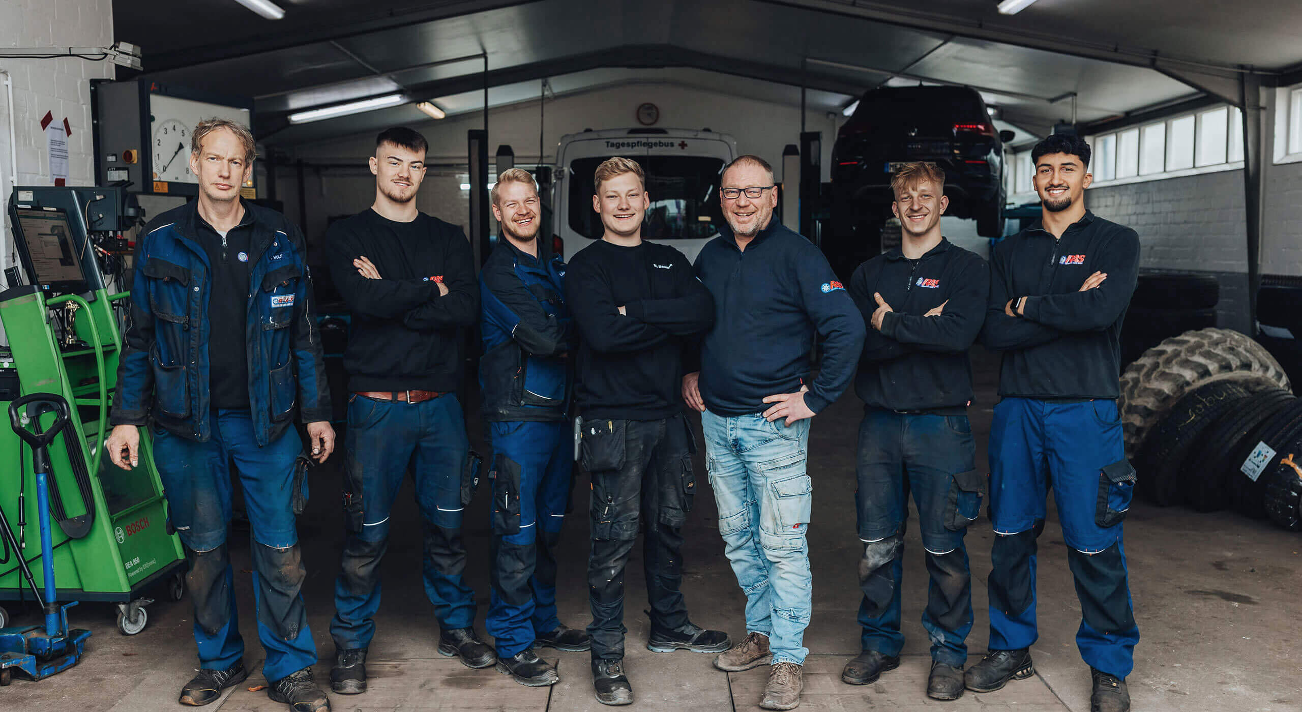 Sechs Personen in Arbeitskleidung und ein Mann in Alltagskleidung stehen lächelnd in einer Kfz-Werkstatt. Im Hintergrund sind Werkzeuge, Autoreifen und ein angehobenes Auto sichtbar. Die Gruppe posiert freundlich für das Foto.