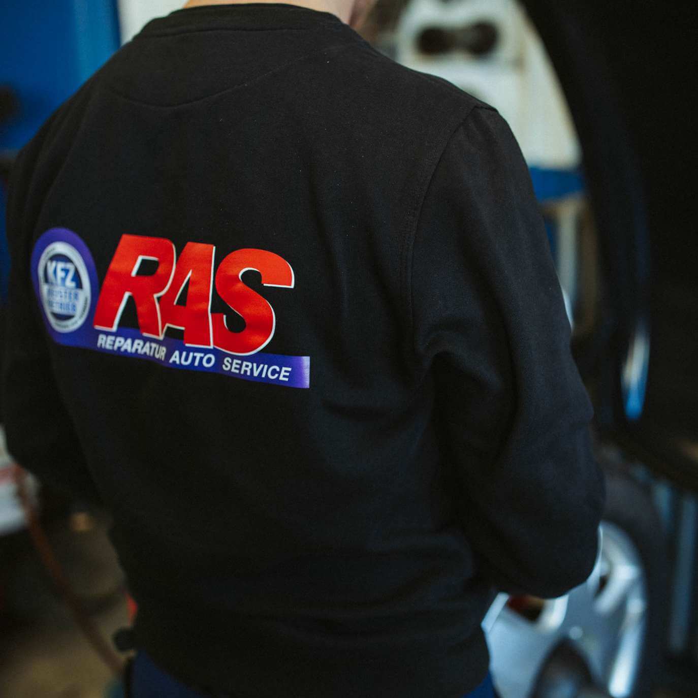 Eine Person in schwarzer Arbeitskleidung steht mit dem Rücken zur Kamera. Auf der Jacke steht in Rot und Blau "RAS" und "Reparatur Auto Service". Im Hintergrund ist unscharf ein Fahrzeug erkennbar.