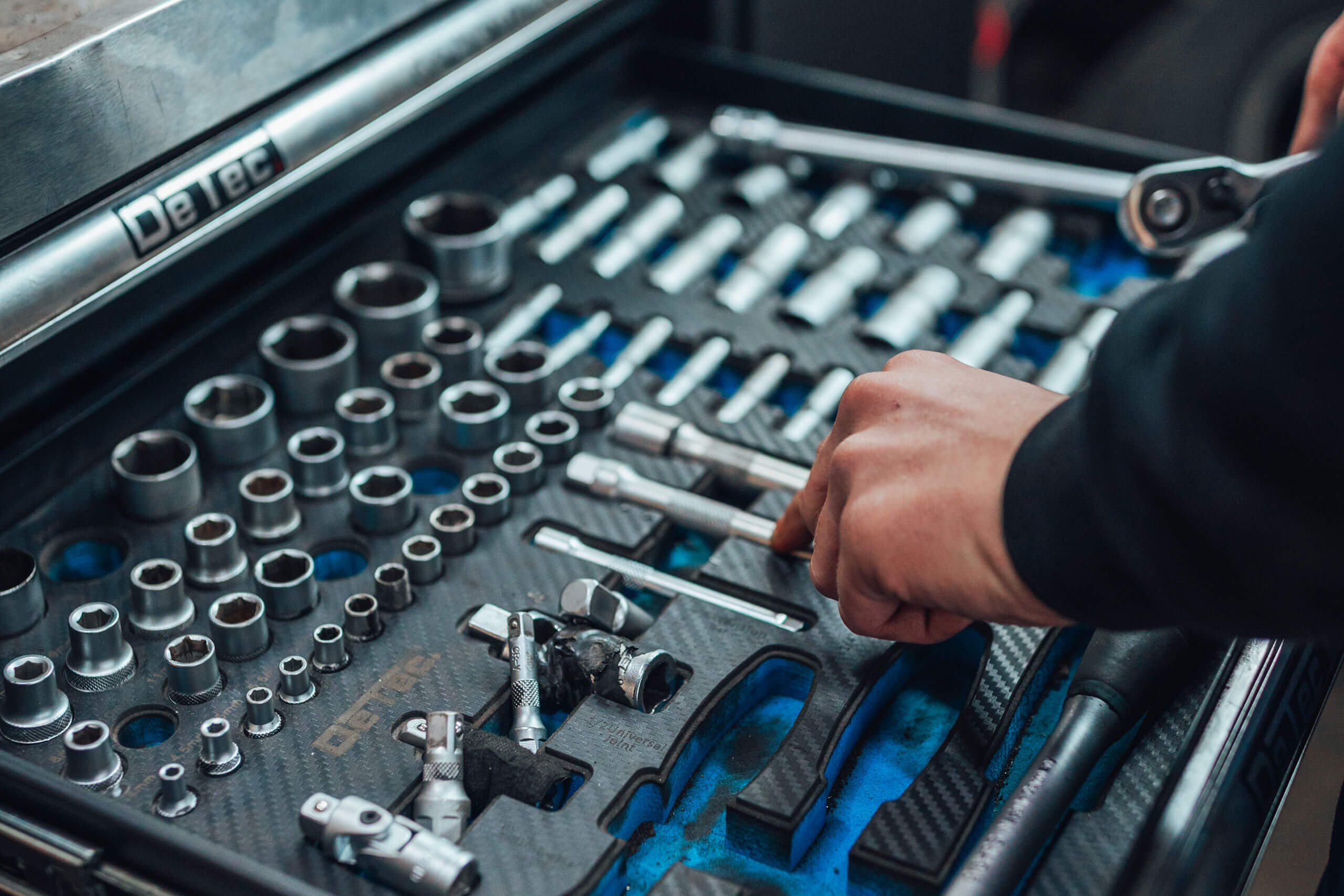 Ein geöffnetes Werkzeugset mit verschiedenen Steckschlüsseln und Ratschengriffen in einer Schublade. Eine Hand greift nach einem der Werkzeuge, die auf einer blauen Matte angeordnet sind.