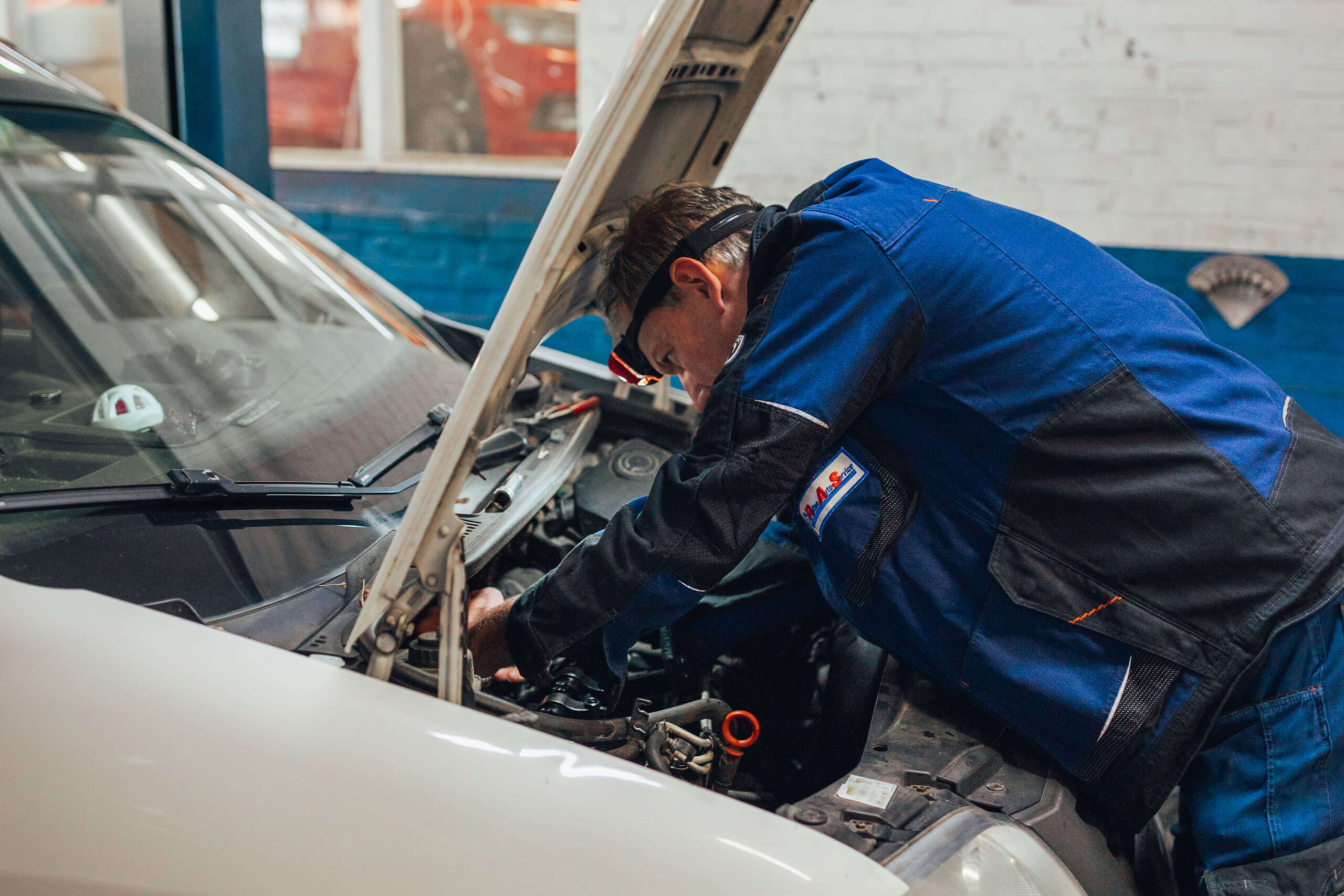 Ein Mechaniker in blauer Arbeitskleidung arbeitet in einer Autowerkstatt am geöffneten Motorraum eines weißen Autos. Er ist nach vorne gebeugt und untersucht die Teile im Motor.